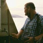Professional charter boat captain sits at the helm of his boat, using a laptop to manage his online bookings and marketing...